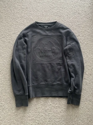Mörkgrå sweatshirt från Superdry - Mörkgrå sweatshirt från Superdry med broderad logga och bergmotiv på bröstet. Tröjan har rund hals, ribbade muddar och är tillverkad i mjukt bomullsmaterial. Perfekt för en avslappnad och stilren look.
