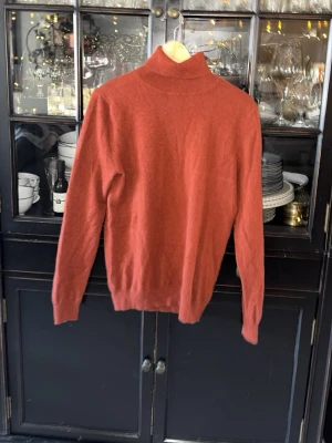 John Henric kaschmir tröja - Jag erbjuder en elegant stickad polotröja i rostfärg med lång ärm och ribbade muddar. Tröjan är försedd med en hög krage och är tillverkad av mjuk kaschmir, vilket gör den idealisk för kyligare väderförhållanden. Den enkla och stilrena designen gör den lämplig för en mängd olika tillfällen.