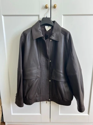 Mörkbrun oversized skinnjacka - Säljer en mörkbrun oversized skinnjacka med bred krage och stora fickor framtill. Jackan har elastisk nederkant och stängs med dragkedja och tryckknappar. 100% lammskinn, foder i viskos. 