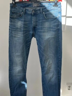Blå slim fit jeans från Dressmann, W33/L32 - Snygga blå slim fit jeans från Dressmann med klassisk femficksmodell och diskreta slitningar framtill. Jeansen har normalhög midja och stängs med knapp och dragkedja. Perfekta för en avslappnad och trendig look.