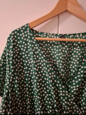 Grön blommig omlottklänning Shein - Grön omlottklänning från Shein med vitt blommönster. Klänningen har korta ärmar, v-ringning och resår i midjan för en bekväm passform. Perfekt för dig som gillar en söt och avslappnad stil.