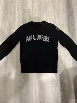 Svart fleece-tröja Parajumpers - Svart fleece-tröja från Parajumpers med stor vit logga över bröstet och patch på ena ärmen. Tröjan har rund halsringning och är tillverkad i mjukt fleecematerial. Perfekt för lager-på-lager och chill dagar.