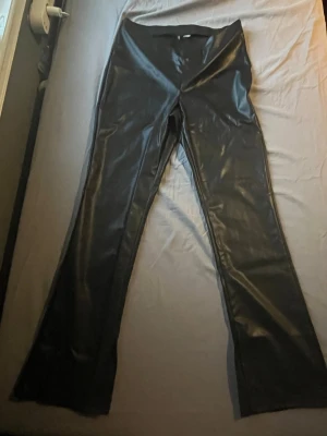 Svarta skinnleggings med utsvängda ben - Snygga svarta leggings i skinnimitation med utsvängda ben och slits på båda benen. Hög midja och stretchigt material som sitter tight och bekvämt. Aldrig använt och har haft dem i fyra år, köptes i fel storlek och vi bytte aldrig dem. Tror dem köptes för 400-500kr.