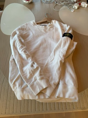 Vit sweatshirt från Stone Island - Vit sweatshirt från Stone Island med klassisk rund hals och ribbade muddar. Ikonisk svart patch med gul logga på vänster ärm. Perfekt för en clean och sliten look. Storlek S. Bra skick. 