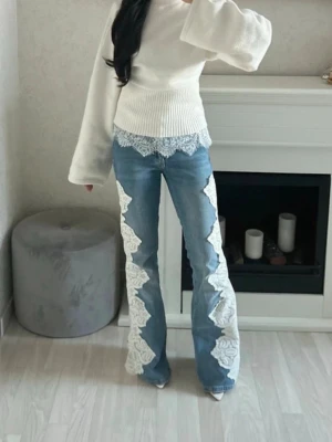 Blå jeans med vit spetsdetalj - Unika blå jeans med breda ben och stora vita spetsapplikationer längs sidorna. Jeansen har hög midja och en snygg, trendig passform som sticker ut. Perfekta för dig som vill ha något annorlunda och coolt i garderoben.