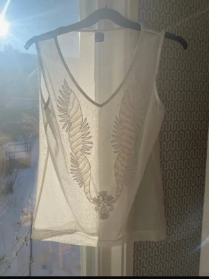 Vit ärmlös linnetopp med broderi - Snygg vit ärmlös linnetopp med v-ringning och broderat fjädermönster framtill. Tunt och lätt material som ger en luftig känsla. Perfekt för varma dagar och enkel att styla till olika looks.