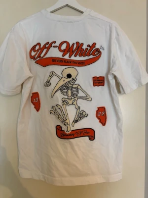 Vit Off-White t-shirt med tryck - Vit t-shirt från Off-White med stort grafiskt tryck på ryggen och mindre tryck framtill. Motivet är ett skelett med orange och svarta detaljer samt texten 'Off-White'. Rund hals och korta ärmar. Perfekt för dig som gillar streetwear.