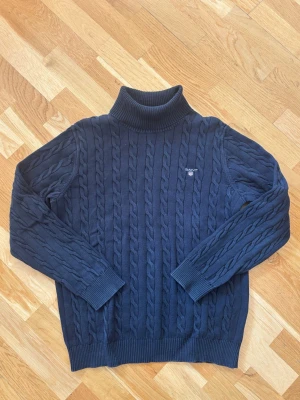 Mörkblå kabelstickad polotröja Gant - Mörkblå kabelstickad polotröja från Gant med broderad logga på bröstet. Tröjan har hög krage, långa ärmar och ribbstickade muddar vid ärmslut och nederkant. Perfekt för lager-på-lager och klassisk stil.