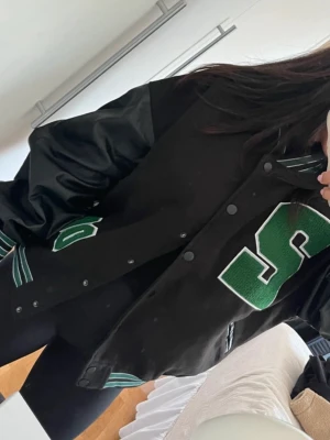 Svart bomberjacka med gröna detaljer - Cool svart bomberjacka med gröna och vita detaljer, stora broderade bokstäver och randiga muddar. Jackan har glansiga ärmar och tryckknappar framtill. Perfekt för en sportig och trendig look. Använd två gånger