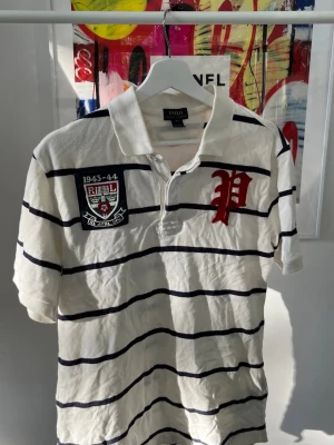 Randig pikétröja från Polo Ralph Lauren - Vit pikétröja från Polo Ralph Lauren med marinblå horisontella ränder, röd broderad bokstav och patch på bröstet. Klassisk krage och korta ärmar. Tillverkad i bomull, perfekt för en avslappnad stil.