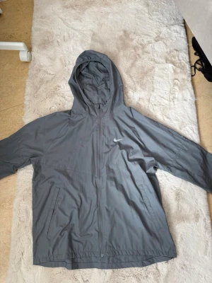 Grå Nike vindjacka med huva - Snygg grå vindjacka från Nike med huva och dragkedja framtill. Jackan har två fickor med dragkedja och diskret Nike-logga på bröstet. Perfekt för löpning eller när det blåser ute.