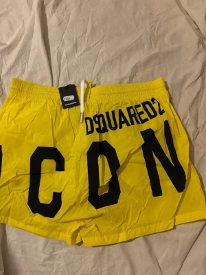 Gula badshorts från Dsquared2 ICON - Snygga gula badshorts från Dsquared2 med stor svart 'ICON'-text och logga på ena benet. De har elastisk midja med vit dragsko och är tillverkade i lätt, snabbtorkande material. Perfekta för stranden eller poolen.