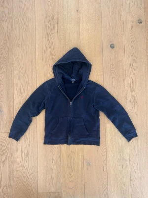 Mörkblå Ralph Lauren zip hoodie - En marinblå Ralph Lauren hoodie med zip, fint skick. Storlek 14-16 år enligt Ralph Lauren, tycker att den är liten i storlek. Kontakta vid minsta fundering! 