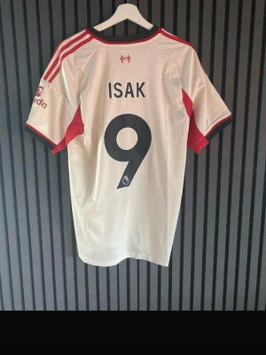 Liverpool bortatröja Isak 9 Adidas - Säljer en vit Liverpool FC bortatröja från Adidas med röda detaljer och tryck. Tröjan har spelarnamnet Isak och nummer 9 på ryggen. Klassisk V-ringning, klubbmärke och sponsorloggor framtill. Perfekt för fotbollsfans!