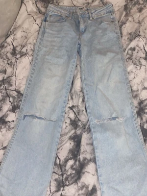 Ljusblå raka jeans med slitningar - Ljusblå jeans från 157 med raka ben och slitna detaljer vid knäna. Klassisk femficksmodell med knapp- och dragkedjegylf. Jeansen har bakfickor med knapp och är tillverkade i denim.