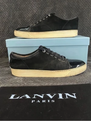 Svarta sneakers från Lanvin - Snygga svarta sneakers från Lanvin med ovandel i mocka och lackad tå. Skorna har vita sulor och klassisk snörning. Skicket är 6/10.Perfekta för dig som gillar stilrena och exklusiva detaljer. Tyvärr så har jag tappat bort boxen så den kommer i en Hugo Boss box istället. Och sulan är lite lös från glansen på skon men kan enkelt fixas hos en sko makare 