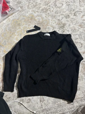 Svart stickad tröja från Stone Island - Snygg svart stickad tröja från Stone Island med klassisk rund halsringning och ikonisk logotyp-patch på ena ärmen. Tröjan har ribbade muddar och är perfekt för dig som gillar stilrena och trendiga plagg.