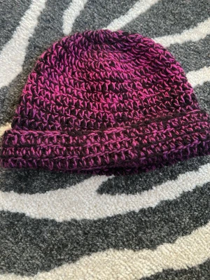Handvirkad lila och svart mössa - virkad mössa i lila och svart garn. Är 22 cm lång för en vikbar kant. Gjord i akrylgarn. Sitter löst på mitt huvud med 55cm i omkrets