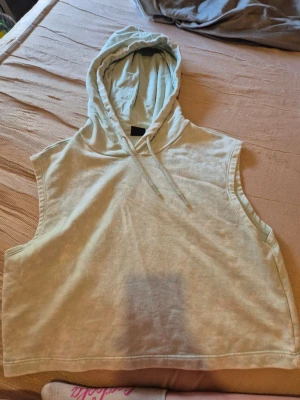 Ärmlös hoodie  - Ljusgrön ärmlös hoodie i mjukt bomullsmaterial. Modellen har huva med dragsko och avslappnad passform, perfekt för en chill och trendig look. Passar till lager-på-lager outfits och ger en sportig vibe.