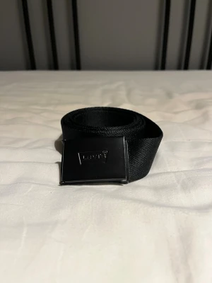 Svart tygbälte från Levi's - Snyggt svart tygbälte från Levi's med stilren metallspänne där loggan är ingraverad. Bältet har en enkel och clean design som passar till de flesta outfits. Perfekt för dig som gillar klassiska accessoarer med en modern touch. Storlek: längd 124cm bredd 3,4cm. OBS! Liten och repa vid slutet av bältet, knappt synbart!