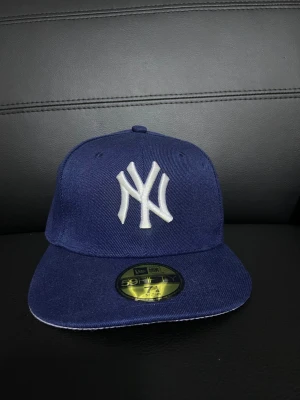 Mörkblå New Era Yankees keps - Snygg mörkblå keps från New Era med klassiska NY Yankees-loggan broderad framtill i vitt. Modellen är 59FIFTY med rak skärm och broderad MLB-logga bak. Perfekt för dig som vill ha en ikonisk streetstyle-accessoar.