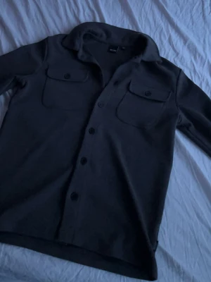 Overshirt - Stilren svart overshirt med knäppning framtill och två bröstfickor med lock och knapp. Overshirten har klassisk krage och raka ärmar. Perfekt att slänga över en t-shirt för en avslappnad look.