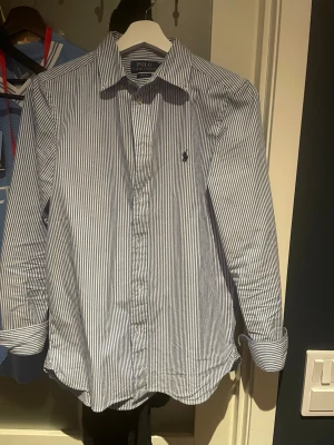 Randig skjorta från Polo Ralph Lauren - Klassisk blå- och vitrandig skjorta från Polo Ralph Lauren med lång ärm och knappar framtill. Skjortan har en broderad logga på bröstet och är tillverkad i bomull. Perfekt för en stilren look.