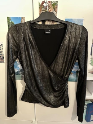 Svart omlottopp i glittrigt tyg - Glittrig svart omlottopp från Gina Tricot med lång ärm och djup v-ringning. Toppens material har en metallic finish som ger en snygg och trendig look. Perfekt för dig som vill sticka ut lite extra.