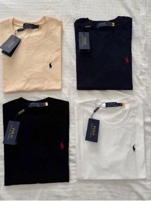 Polo Ralph Lauren t-shirts i fyra färger - Fyra klassiska t-shirts från Polo Ralph Lauren i färgerna beige, marinblå, svart och vit. Alla har rund hals, kort ärm och den ikoniska broderade loggan på bröstet. Perfekta basplagg i mjuk bomull för en stilren look.