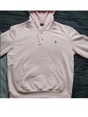 Ljusrosa hoodie från Polo Ralph Lauren - Ljusrosa hoodie från Polo Ralph Lauren med klassisk huva och dragsnören. Ikonisk blå logga broderad på bröstet och stor magficka framtill. Mjuk bomullskänsla och ribbade muddar vid ärmslut och nederkant. Perfekt för en chill look. Finns ett litet hål vid fickan, kontakta för bild