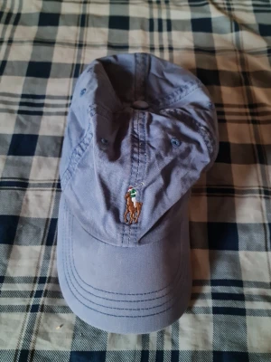 Ljusblå keps från Polo Ralph Lauren - Snygg ljusblå keps från Polo Ralph Lauren med den klassiska broderade björnen framtill. Kepsen har böjd skärm och justerbar rem baktill. Perfekt accessoar för en avslappnad och sportig stil.
