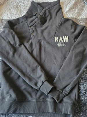 Mörkgrå RAW hoodie med hög krage - Snygg mörkgrå hoodie från RAW med hög krage och tryck på bröstet. Hoodien har ribbade muddar vid ärmslut och nederkant samt knappdetaljer vid halsen. Tillverkad i mjukt bomullsmaterial, perfekt för en avslappnad streetstyle.
