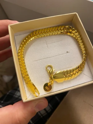 18K Guldpläterad armband  - Snyggt armband i guldfärgad metall med platt, bred kedja och karbinlås. Märkt med en liten bricka där det står 18K. Perfekt accessoar för dig som gillar stilrena och trendiga smycken.