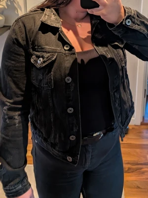 Svart jeansjacka med slitningar - Snygg svart jeansjacka med coola slitningar och silverfärgade knappar. Klassisk modell med två bröstfickor med lock och knapp. Jackan har en rak passform och är tillverkad i jeansmaterial. Perfekt för dig som gillar en edgy stil.