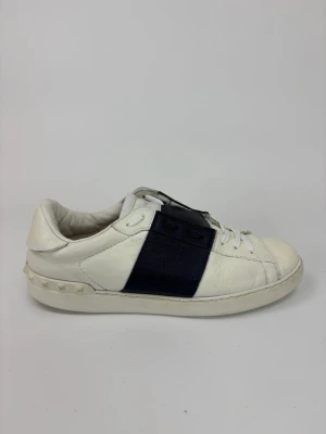 Valentino vita sneakers med svart detalj - Snygga vita sneakers från Valentino med en bred svart läderdetalj över snörningen och nitar på hälen. Skorna har en klassisk låg profil och är tillverkade i läder med vita snören och sula. Perfekta för en stilren och trendig look.