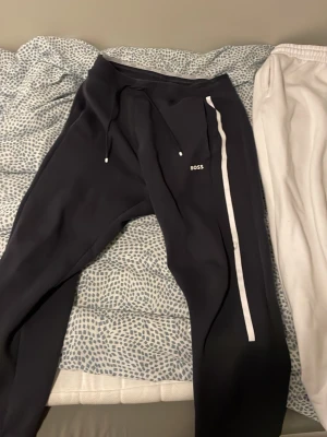 Svarta mjukisbyxor från BOSS - Svarta mjukisbyxor från BOSS med vit rand längs benet och dragsko i midjan. Snygg och enkel design med BOSS-logga på låret. Perfekta för en chill och sportig stil.