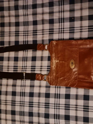 Brun axelväska i läder - Snygg brun axelväska i äkta läder med justerbar svart axelrem och guldfärgat vridlås framtill. Väskan har en klassisk rektangulär form och en framficka för extra förvaring. Perfekt för dig som gillar stilrena accessoarer.