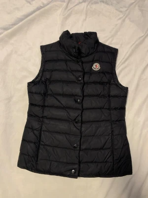 Moncler väst - Svart moncler bäst med slim fit modell, som man ser på sista bilden så är knappen längst ner sönder