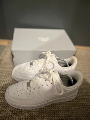 Nike Air Force 1 vita sneakers - Klassiska Nike Air Force 1 sneakers i helvitt läder med perforerad tå, vit sula och vita skosnören. Ikonisk siluett med låg profil och diskret Nike-logga på sidan. Perfekta för en clean och stilren look. Använda två gånger & säljer pågrund av stor storlek.