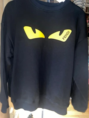 Svart Fendi sweatshirt med gula ögon - Oanvänd svart sweatshirt från Fendi med ikoniska gula monsterögon framtill och rund hals. Säljs, har ej kommit till användning. Storlek S, funkar i M även. Stilrent, passar med olika stilar