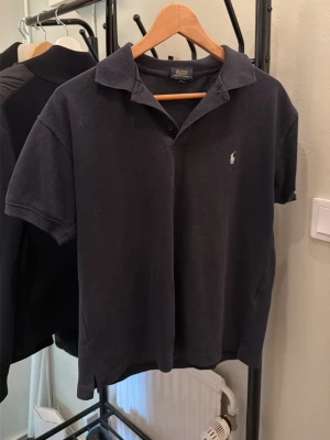 Svart pikétröja från Ralph Lauren - Klassisk svart pikétröja från Ralph Lauren med korta ärmar och krage. Storlek S men sitter som xs skulle säga den passar folk mellan 160-170 cm. Tröjan har en liten broderad logotyp på bröstet och är tillverkad i mjuk bomull. Perfekt för en stilren och avslappnad look. 