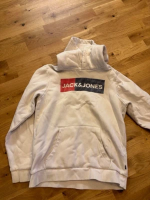 Vit hoodie från Jack & Jones - Snygg vit hoodie från Jack & Jones med stor logga i rött och blått framtill. Klassisk känguruficka och huva. Perfekt för en chill och avslappnad stil. Mjuk bomullskänsla och ribbade muddar vid ärmslut och nederkant.