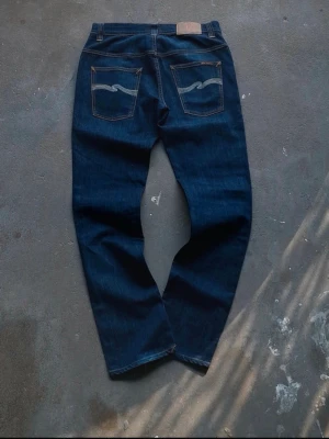 Thinn Finn Nudie Jeans - Säljer ett par mörkblå Nudie Jeans Co jeans i mycket fint skick. Klassisk rådenim-look med orange kontrastsömmar och den ikoniska vågstickningen på bakfickorna.  • Storlek: W33/L32 • Färg: Mörk indigo/blå • Skick: Väldigt bra skick, minimalt använda • Modell med rak/slim passform • Äkta läderpatch och originaldetaljer kvar  Perfekta för både vardag och mer uppstyrd stil. Snygga fades börjar komma fram och jeansen kommer bara bli finare med användning.  Kan skickas eller möt