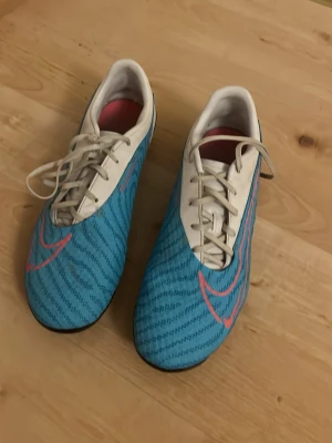 Nike blå fotbollsskor med dobbar - Säljer ett par Nike fotbollsskor i blått med vita och rosa detaljer. Skorna har snörning, svart yttersula med dobbar och en stor rosa Nike-logga på sidan. Ovandelen är i syntetmaterial och insidan är vit med rosa detaljer. Perfekta för spel på gräsplan.