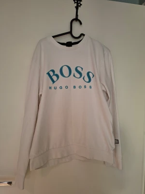 Vit sweatshirt från Hugo Boss - Vit sweatshirt från Hugo Boss med stort blått BOSS-tryck och HUGO BOSS-text på bröstet. Tröjan har rund halsringning, långa ärmar och en relaxed fit. Perfekt för en chill och stilren look. Materialet känns mjukt och bekvämt mot huden.