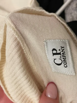 Vit stickad tröja från C.P. Company - Snygg vit stickad tröja från C.P. Company med hög krage och ribbade muddar. Passar både killar och tjejer är mysig på vintern och sommar kväll, Tröjan har en ikonisk detalj med svart lins på ärmen. Perfekt för dig som gillar stilrena och trendiga plagg. 