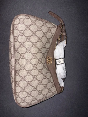 Beige GG canvas kuvertväska från Gucci - Snygg kuvertväska från Gucci i beige canvas med klassiskt GG-mönster och bruna läderdetaljer. Väskan har guldfärgad logga och dragkedja upptill samt en smal handledsrem. Perfekt för dig som gillar lyxiga accessoarer med ikonisk design.