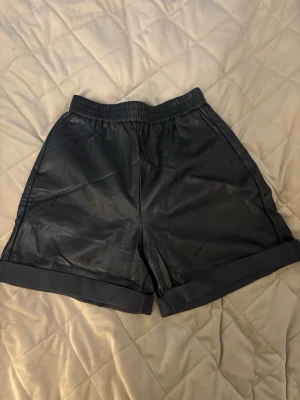 Svarta shorts i skinnimitation - Snygga svarta shorts i skinnimitation med elastisk midja och uppvikta benslut eller långa. Skrynkliga pågrund av att jag vikt upp dem.