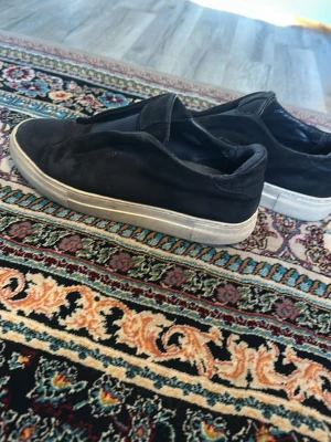 Svarta slip-on sneakers från Axel Arigato  - Stilrena svarta slip-on sneakers från Axel Arigato med vit sula. Skorna har en enkel och clean design utan snörning, vilket gör dem lätta att ta på. Ovandelen är i mockaliknande material och sulan är i gummi. Perfekta för en avslappnad och trendig look. Med en bra läder.