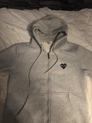 Comme des Garçons | Zip - Säljer en grå zip-hoodie från Comme des Garçons Play med svart hjärta och ögon på bröstet samt stort svart hjärta med ögon på ryggen. Hoodien har dragkedja, fickor fram och justerbar huva. Tillverkad i mjukt bomullsmaterial, perfekt för en chill och stilren look.